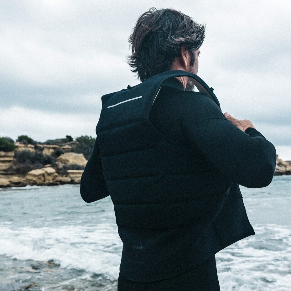 Riders enfilant la veste de protection MANERA Vagabond Impact 2025, face à une mer agitée et une falaise sous un ciel nuageux – idéale pour le wingfoil et le downwind